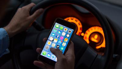 Ein Mann sitzt in einem Auto am Steuer und hält ein Smartphone in der Hand.