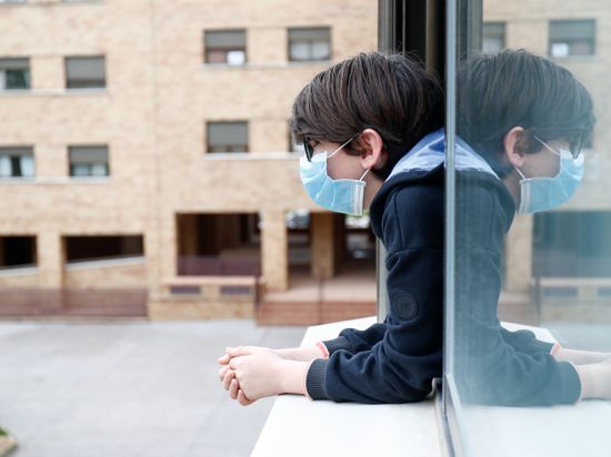 Ein Kind mit Mundschutz schaut aus dem Fenster seines Hauses. 