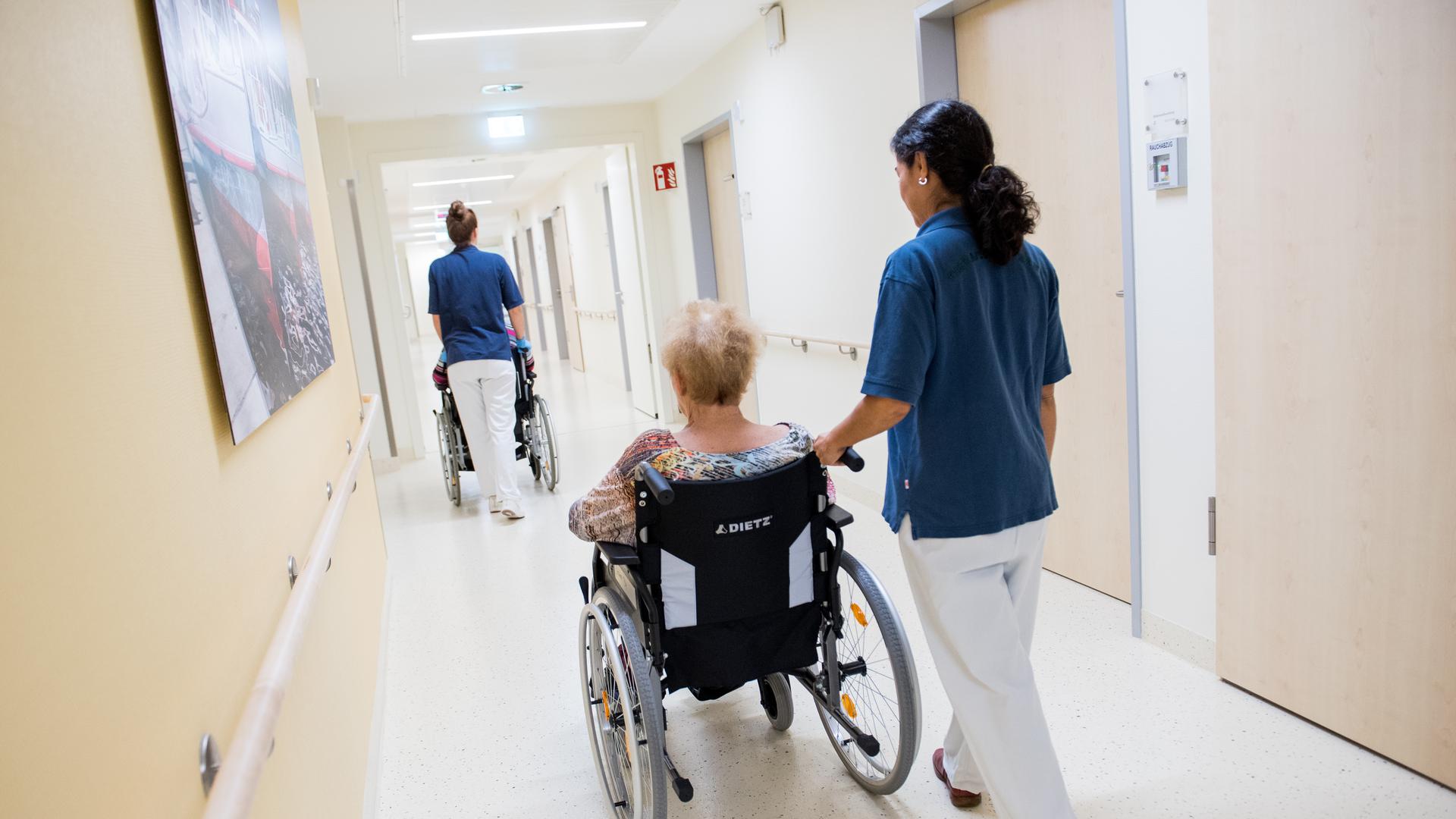 Zwei Pflegerinnen schieben Patienten mit Rollstühlen auf einem Flur in einem Krankenhaus.