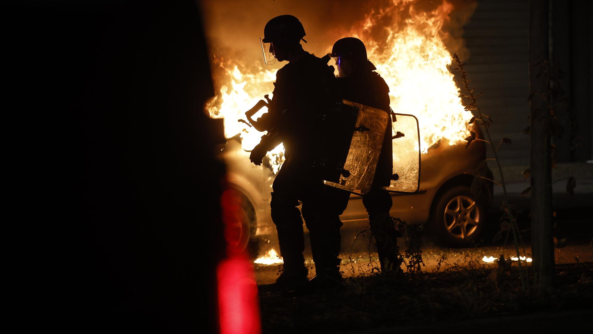 Bereitschaftspolizisten stehen in der Nähe eines brennenden Autos im Viertel La Meinau. Mit Einschränkungen des öffentlichen Lebens will die Regierung in Frankreich die schweren Krawalle nach dem Tod eines Jugendlichen durch einen Polizeischuss eindämmen. +++ dpa-Bildfunk +++