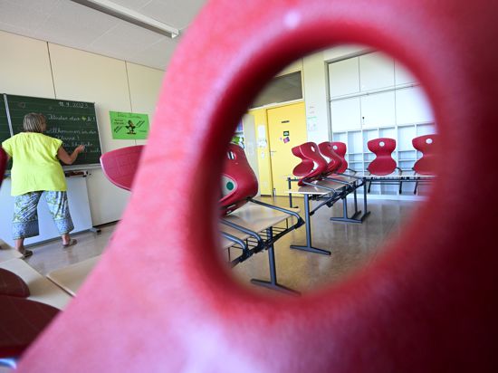 Stühle sind in einem Klassenzimmer einer Grundschule in Stuttgart auf Tische aufgereiht, im Hintergrund bereitet eine Lehrerin einen Tafelanschrieb vor. 