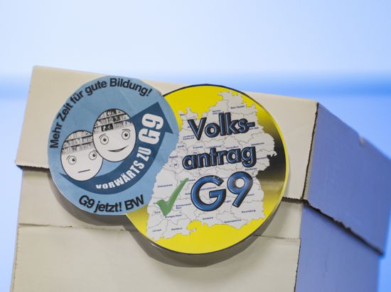 Ein Karton steht auf einem Tisch, daran zwei Aufkleber mit der Aufschrift „Vorwärzt zu G9“ und „Volksantrag G9“. 