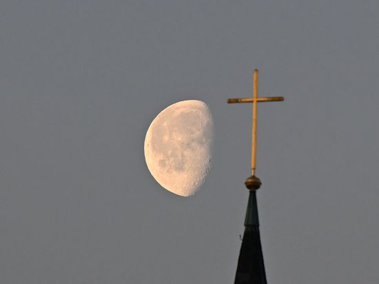 Der Mond steht im Morgenlicht hinter einem Kreuz auf einem Kirchturm. 