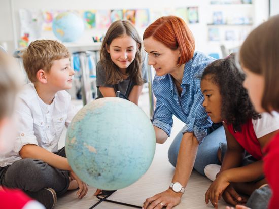 A group of small school kids with unrecognizable teacher sitting on the floor in class, learning geography.