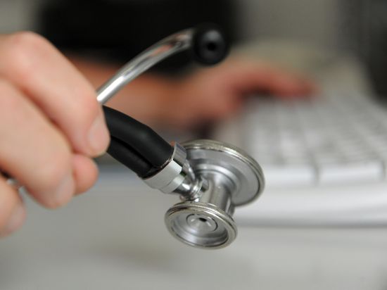 Ein Arzt hält in einem Behandlungszimmer in seiner Praxis ein Stethoskop in der Hand, mit der anderen Hand bedient er eine Computertastatur.