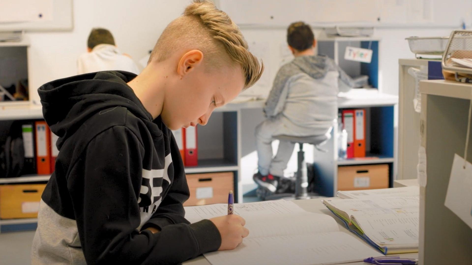 Kind schreibt an einem Schreibtisch in ein Arbeitsheft. Im Hintergrund andere Schüler an Schreibtischen mit Aktenordner-Regalen.
