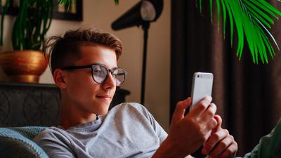 Generation Z concept. Smiling boy in glasses using smartphone sitting in the chair indoor. 