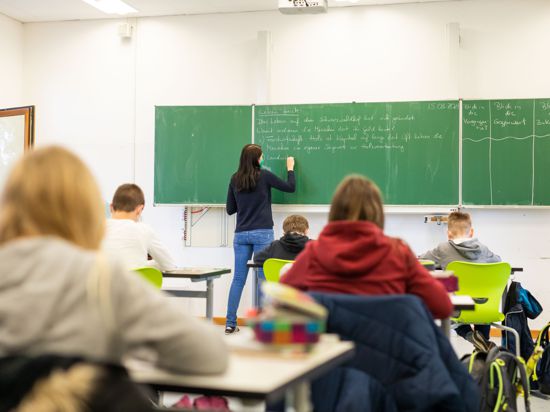 Schülerinnen und Schüler einer fünften Klasse der Johanniter Realschule Heitersheim sitzen während des Unterrichts in ihrem Klassenzimmer. Im Hintergrund schreibt eine Lehrerin an die Tafel.