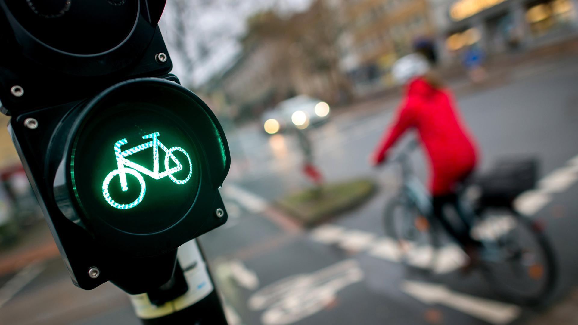 Eine grüne Ampel mit Fahrradsymbol, im Hintergrund ein Fahrradfahrer mit roter Jacke und Helm.