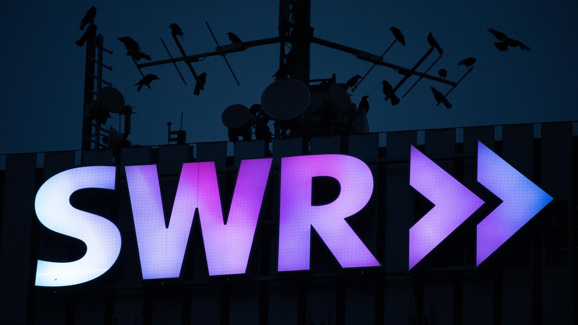 Das Logo des Südwestrundfunks (SWR) leuchtet am frühen Morgen am Funkhaus, während Vögel auf den Antennen am Dach sitzen. 