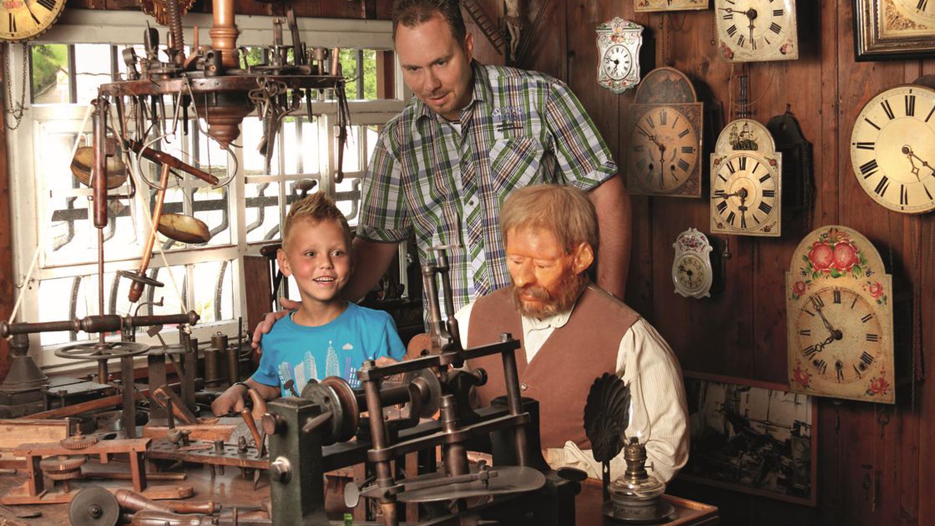 Ein Vater und sein Sohn stehen neben einer lebensgroßen Uhrmacher-Puppe in einer Uhrmacherwerkstatt.