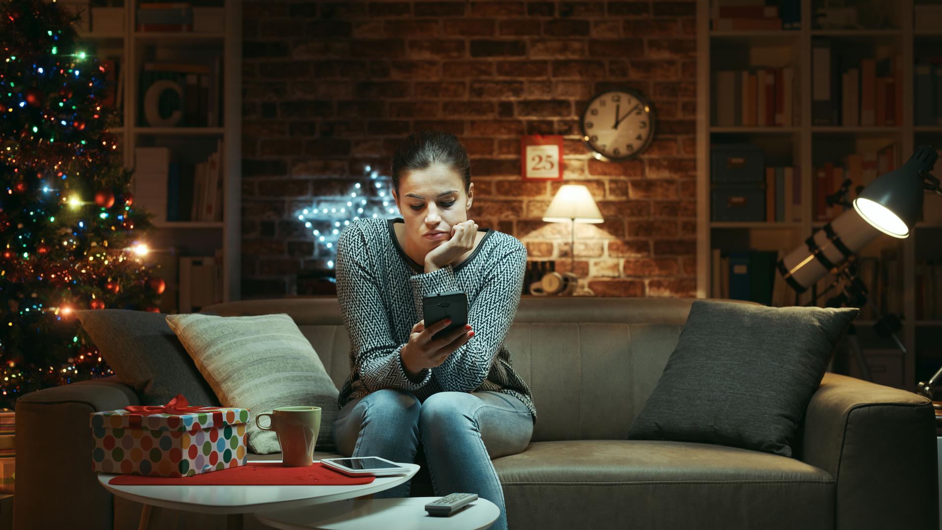 Sad woman celebrating Christmas at home alone: she is sitting on the sofa and waiting for messages on her smartphone