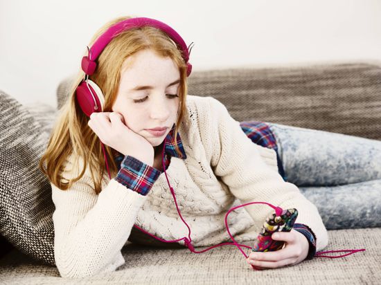 Ein Mädchen sitzt mit Kopfhörern auf einer Couch und hört sich etwas an.