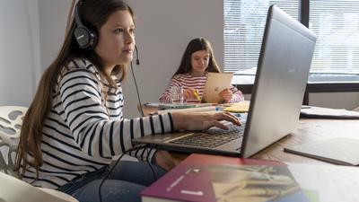 Zwei Schülerinnen sitzen an einem Schreibtisch und lernen mit Laptop und Tablet.