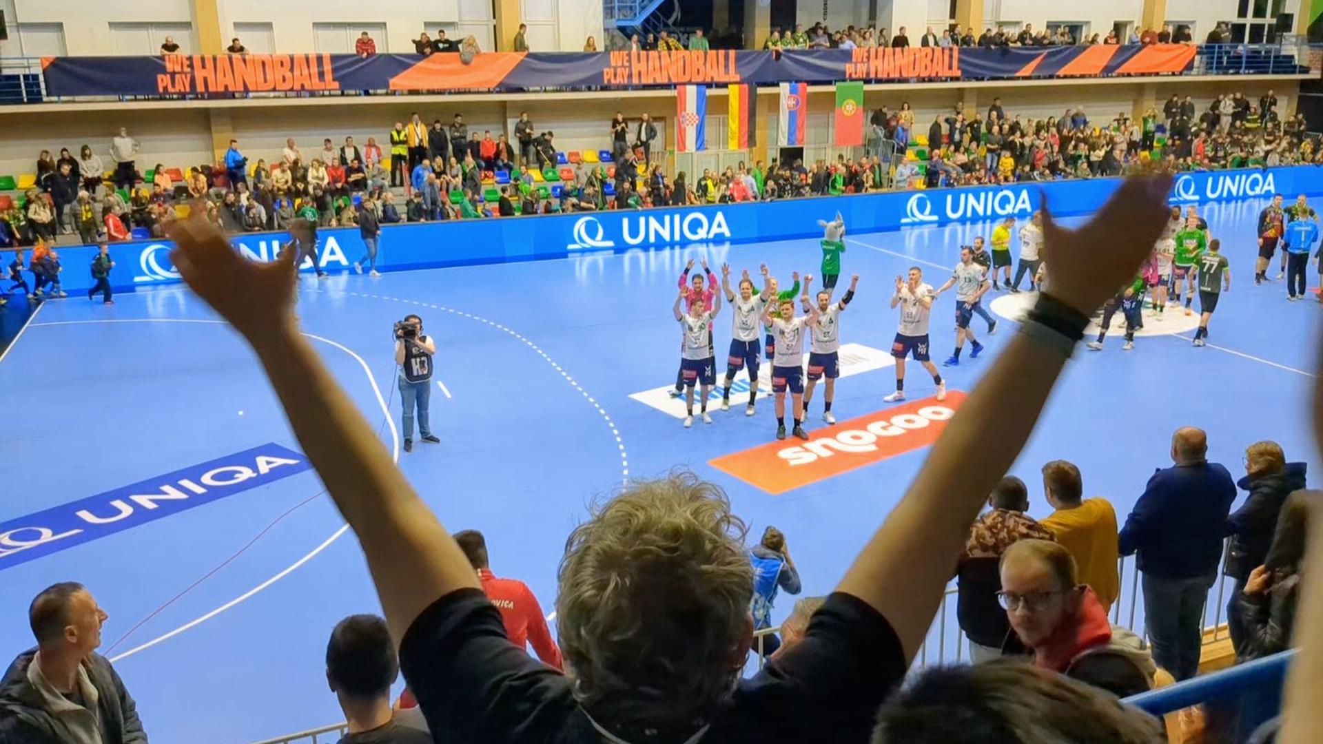 Mann steht mit dem Rücken zur Kamera, beide Hände in die Luft gestreckt, im Hintergrund eine Handballmannschaft.