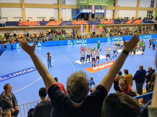 Mann steht mit dem Rücken zur Kamera, beide Hände in die Luft gestreckt, im Hintergrund eine Handballmannschaft.