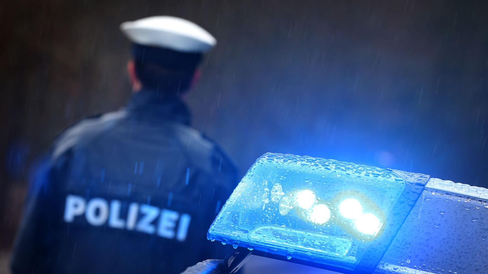 Ein Polizist steht im Regen vor einem Streifenwagen dessen Blaulicht aktiviert ist.