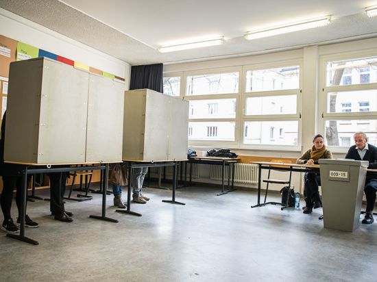 Ehrenamtliche Wahlhelfer sitzen in einem Wahllokal vor einer Urne.
