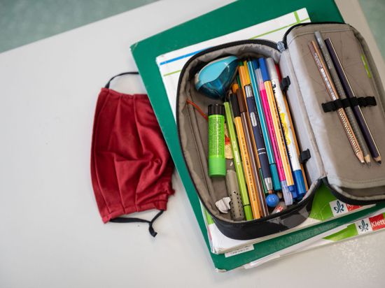 Eine Mund-Nasen-Bedeckung liegt neben einem Mäppchen und Schulbüchern auf einem Schultisch.