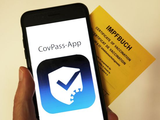 Das Logo der CovPass-App ist neben einem Impfbuch auf einem Smartphone zu sehen.