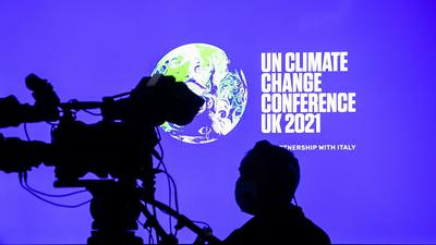 Ein Kameramann steht bei der Weltklimakonferenz in Glasgow vor einem Bildschirm, der das Logo der Konferenz zeigt.