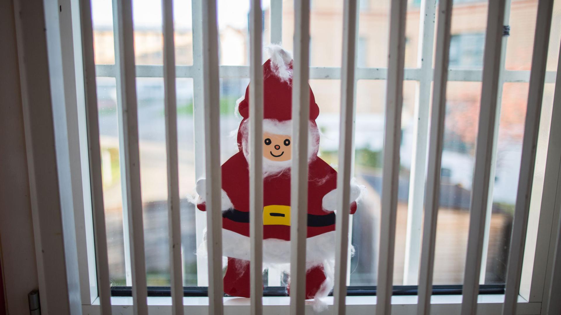 Ein gebastelter Weihnachtsmann klebt hinter einem Gitter einer Justizvollzugsanstalt an der Fensterscheibe.
