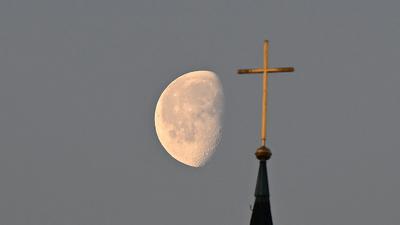 Der Mond steht im Morgenlicht hinter einem Kreuz auf einem Kirchturm.