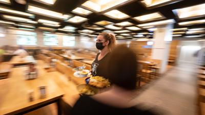 Eine Angestellte mit Mund-Nasen-Schutz trägt in einem gastronomischen Betrieb in der Innenstadt Speisen an einen Tisch.