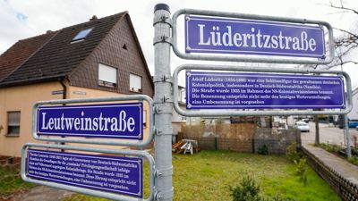 Straßenschilder mit der Aufschrift „Leutweinstraße“ und „Lüderitzstraße“ sowie Schilder zu Theodor Leutwein und Adolf Lüderitz mit Begleittexten stehen an einer Straßenkreuzung.