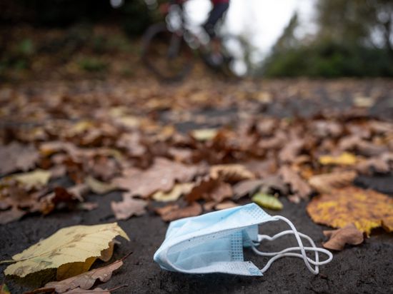 Eine weggeworfene Maske liegt zwischen Herbstlaub auf einer Straße, während ein Radfahrer vorbeifährt.