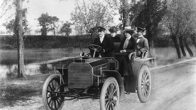 In einem Mercedes sind im frühen 20. Jahrhundert zwei Herren und zwei Damen auf einer Landstraße unterwegs.