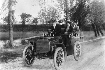 In einem Mercedes sind im frühen 20. Jahrhundert zwei Herren und zwei Damen auf einer Landstraße unterwegs.