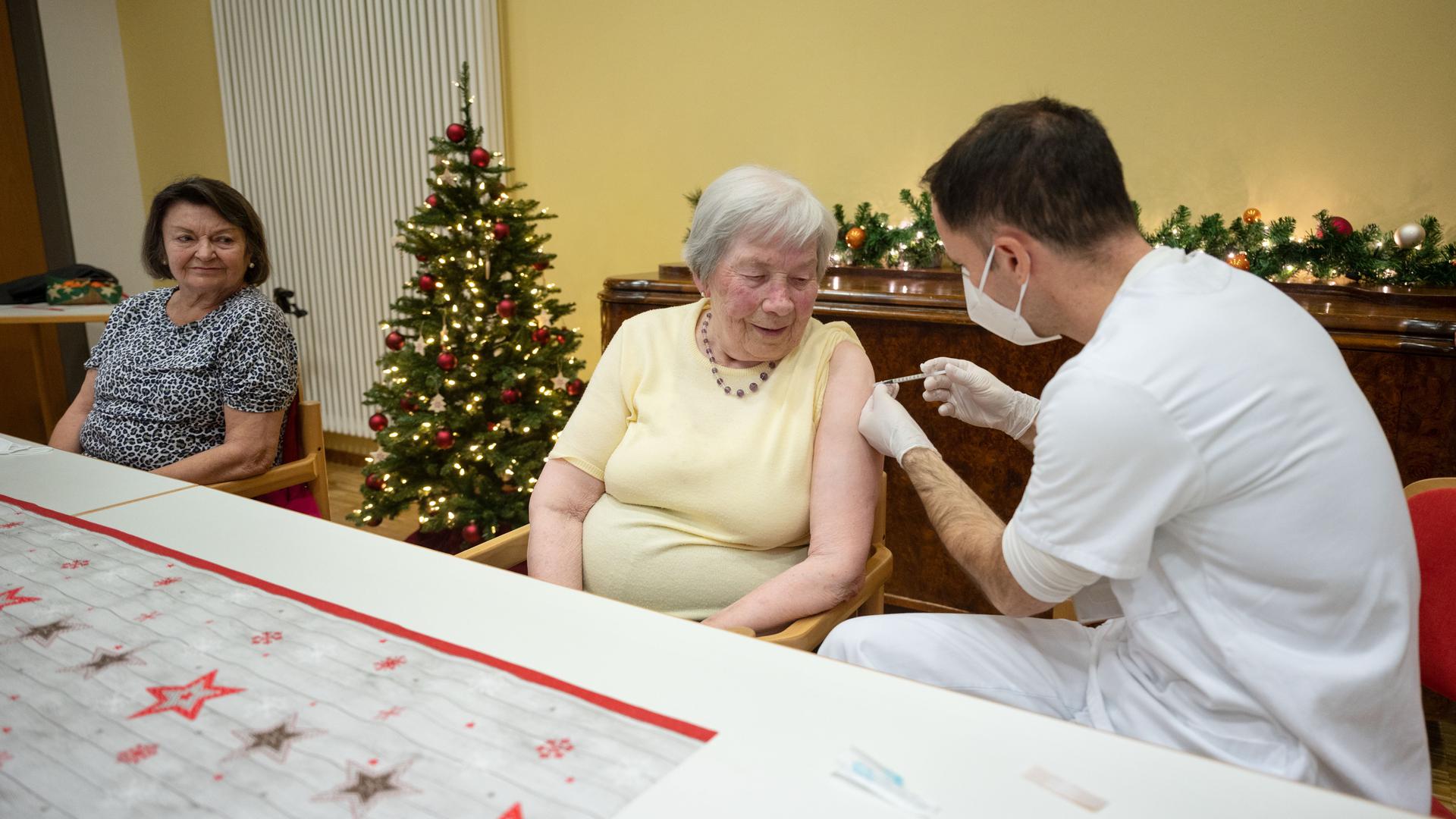 Eine 86 Jahre alte Frau (M) wird in einem Altenpflegeheim von einem Mitglied eines Mobilen Impfteams des Klinikums Stuttgart mit einer Dosis eines Covid-19 Impfstoffes geimpft, daneben sitzt eine 64 Jahre alte Frau, die auf ihre Impfung wartet. Am Sonntag haben die Corona-Impfungen mit dem Impfstoff von Biontech/Pfizer in Deutschland begonnen.