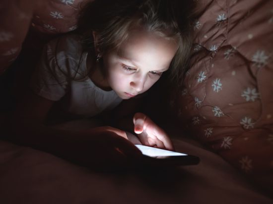 Ein Mädchen schaut unter der Bettdecke auf ihr Smartphone, das im Dunkeln leuchtet.