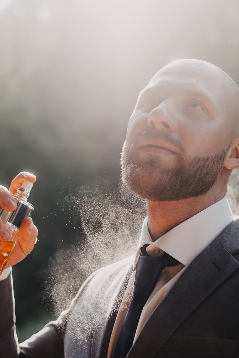 Ein Mann mit Bart steht in der Natur und besprüht sich mit einem Parfum, Duftnebelumwölkt ihn.