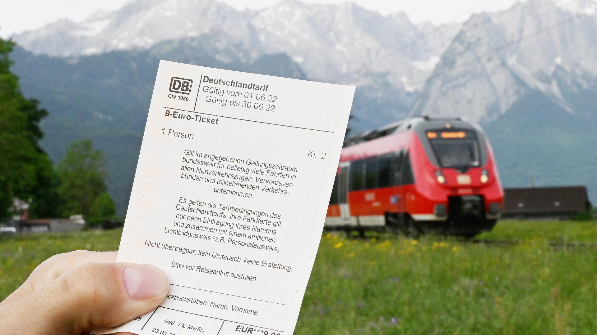 Garmisch-Partenkirchen: Ein Mann hält ein 9-Euro-Ticket in der Hand, im Hintergrund fährt ein Zug der Deutschen Bahn vor der Bergkulisse des Wettersteingebirges vorbei.