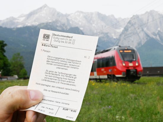 Garmisch-Partenkirchen: Ein Mann hält ein 9-Euro-Ticket in der Hand, im Hintergrund fährt ein Zug der Deutschen Bahn vor der Bergkulisse des Wettersteingebirges vorbei.