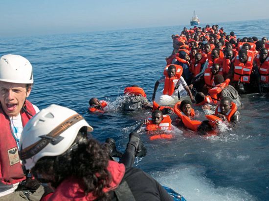 Ein Seerettungsboot rettet Flüchtlinge aus einem kleinen Boot. Die Flüchtlinge stürzen sich aus dem Schlauchboot ins Wasser.