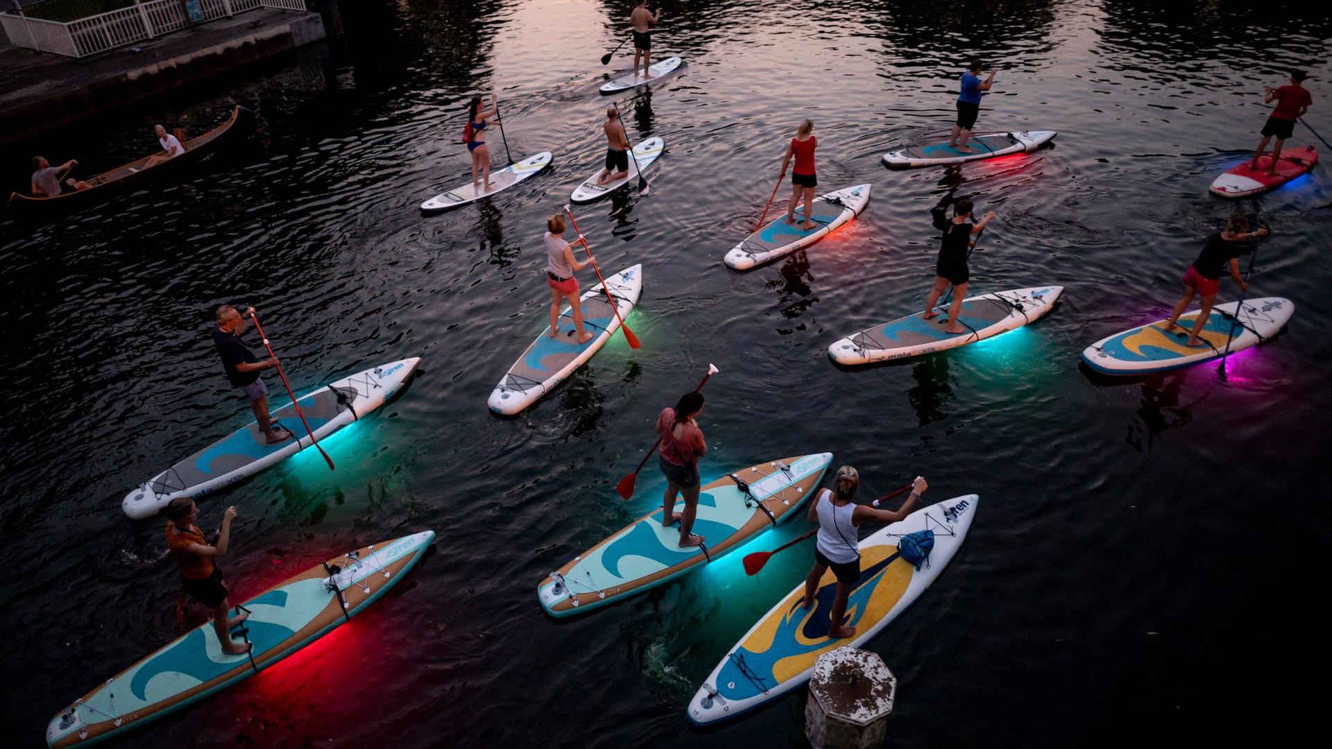 Teilnehmer einer Ausflugsgruppe fahren mit leuchtenden LED-Stand-Up-Paddles in den Abendstunden über einen Alsterfleet in Hamburg.