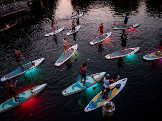 Teilnehmer einer Ausflugsgruppe fahren mit leuchtenden LED-Stand-Up-Paddles in den Abendstunden über einen Alsterfleet in Hamburg.