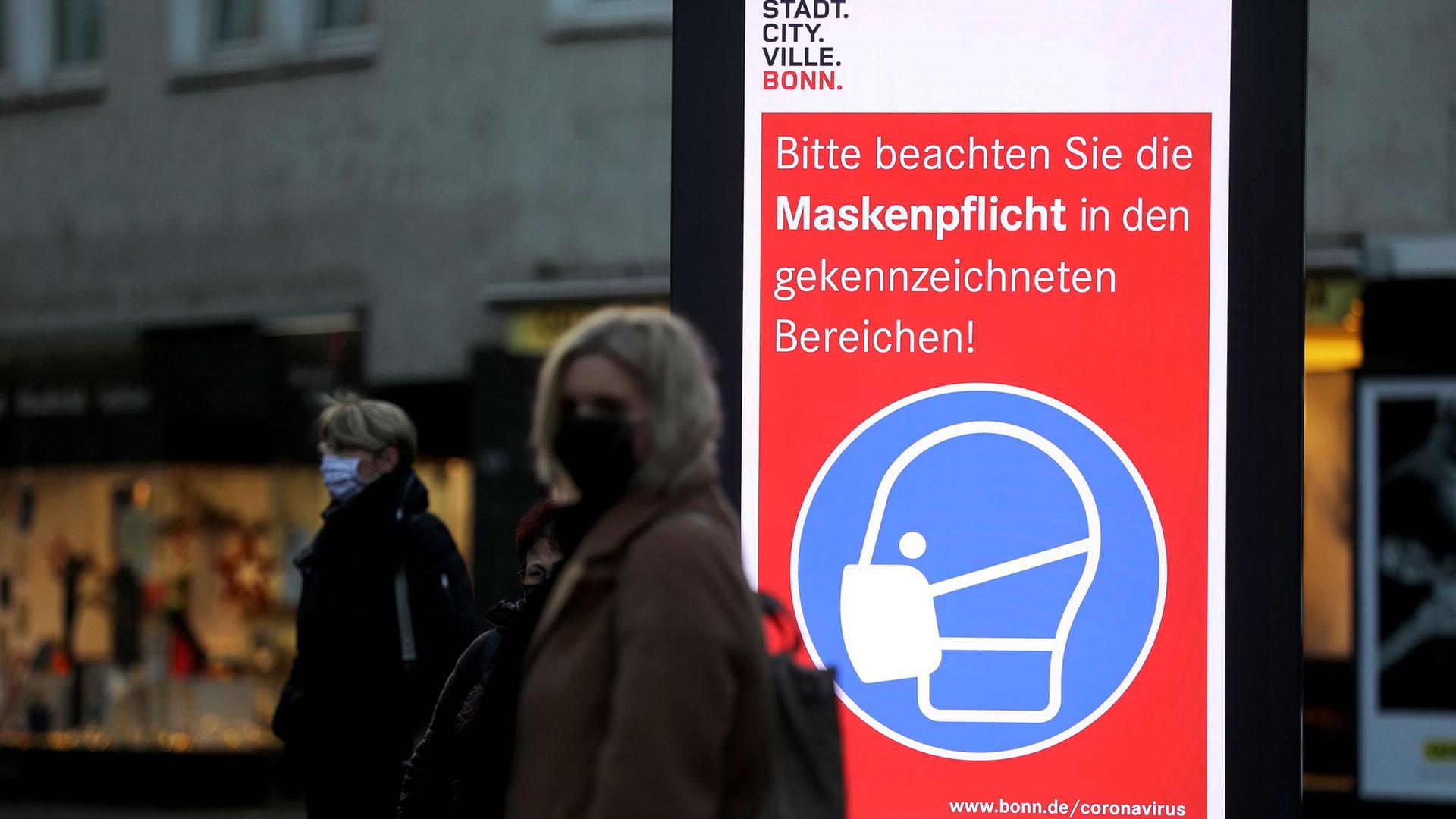 Frauen stehen neben einem Hinweisschild zur Maskenpflicht an einer Haltestelle in Bonn. Die Länder fordern vor den Beratungen mit Kanzlerin Merkel strengere Kontaktbeschränkungen, um die hohen Coronazahlen zu senken.