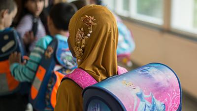 Das Symbolfoto zeigt ein Mädchen mit Kopftuch vor einem Klassenzimmer.