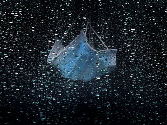 Ein medizinischer Mundschutz hängt am Rückspiegel eines im Regen parkenden Autos.
