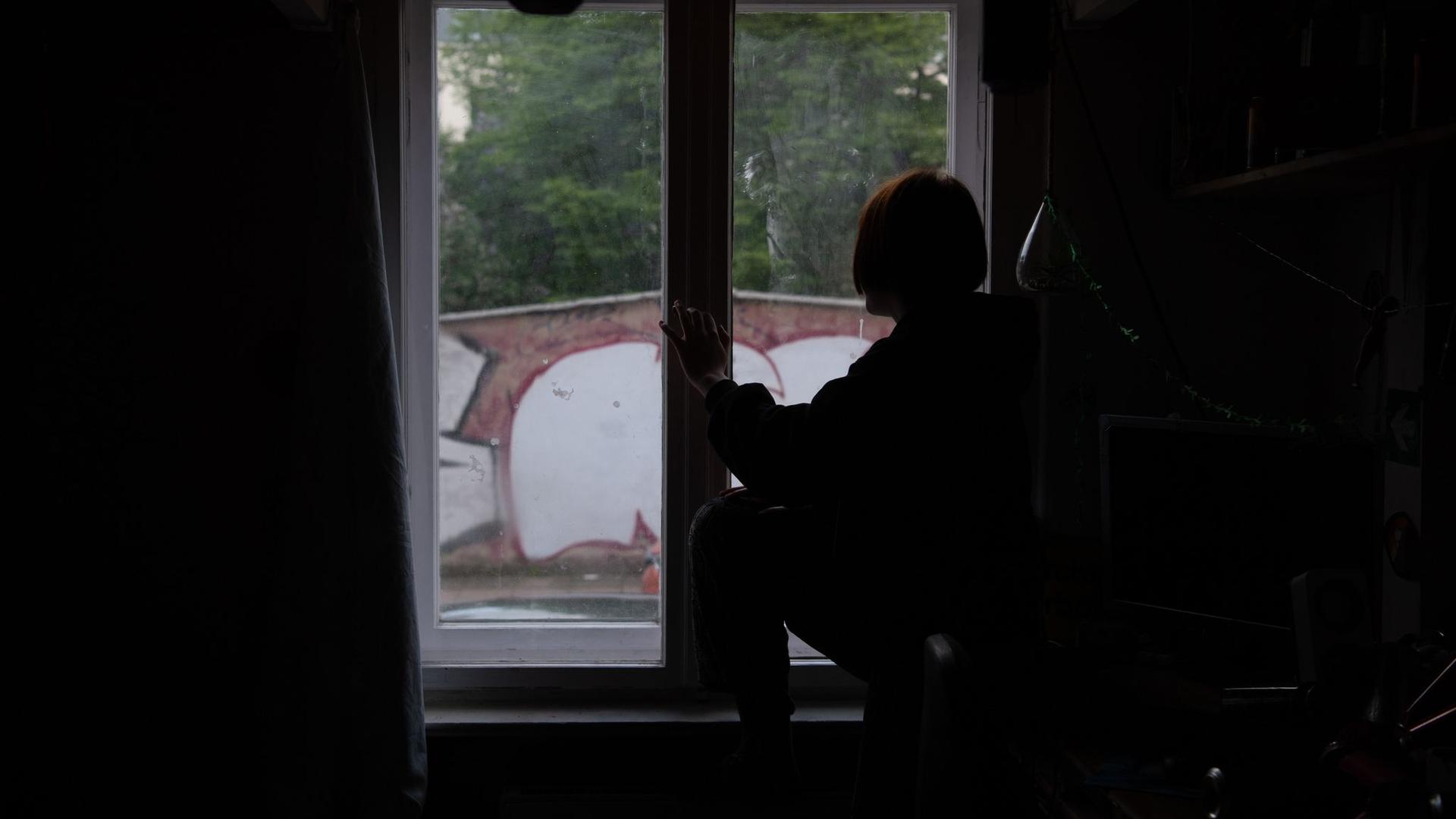 Eine Jugendliche sitzt in einem abgedunkelten Zimmer am Fenster.