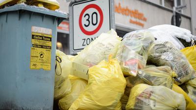 Mülltonnen und gelbe Säcke mit Kunststoff-Abfällen stapeln sich auf einem Fußweg.