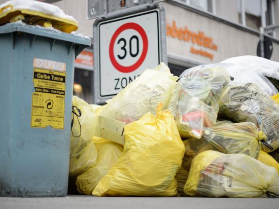 Mülltonnen und gelbe Säcke mit Kunststoff-Abfällen stapeln sich auf einem Fußweg.