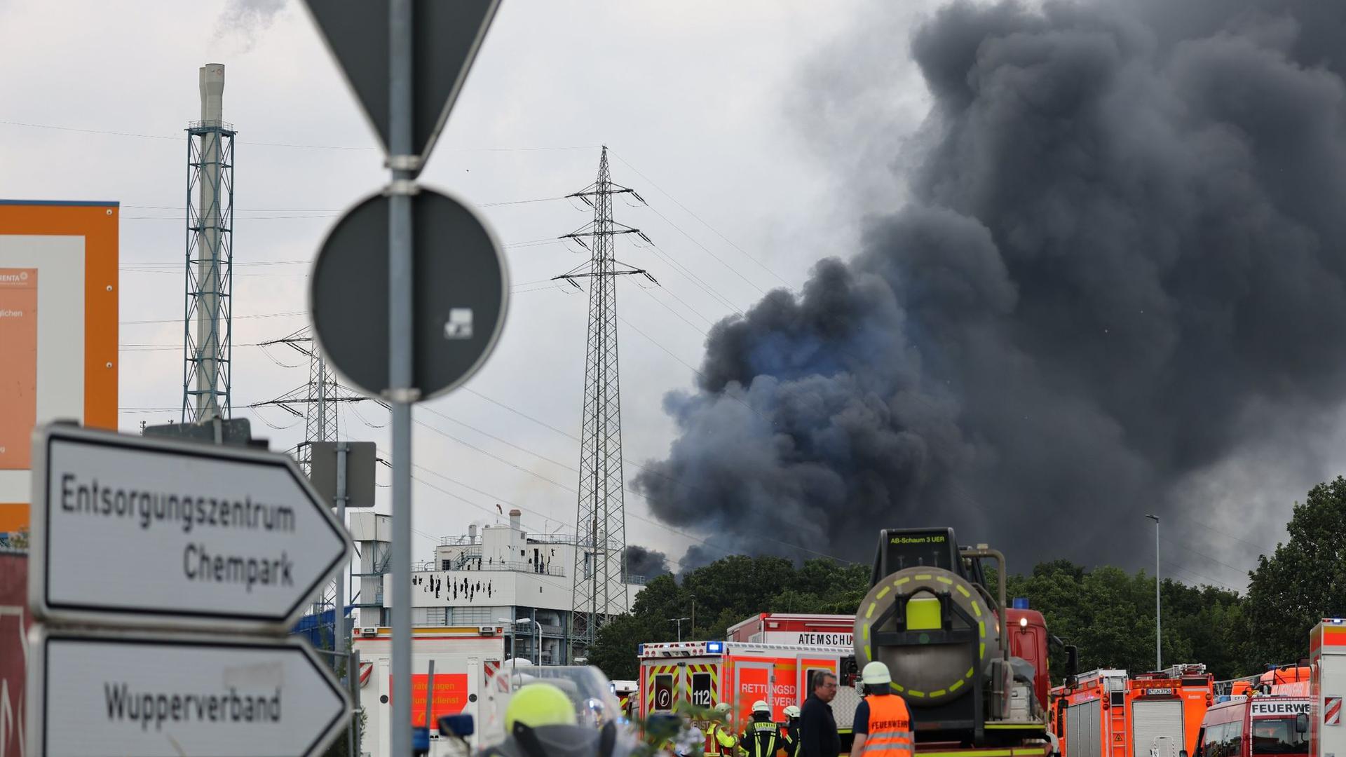 Einsatzfahrzeuge der Feuerwehr und  Rettungsdienste unweit einer Zufahrt zum Chempark über dem eine dunkle Rauchwolke aufsteigt. Etwa eine Woche nach der Explosion in Leverkusen ist ein weiteres Todesopfer geborgen worden.