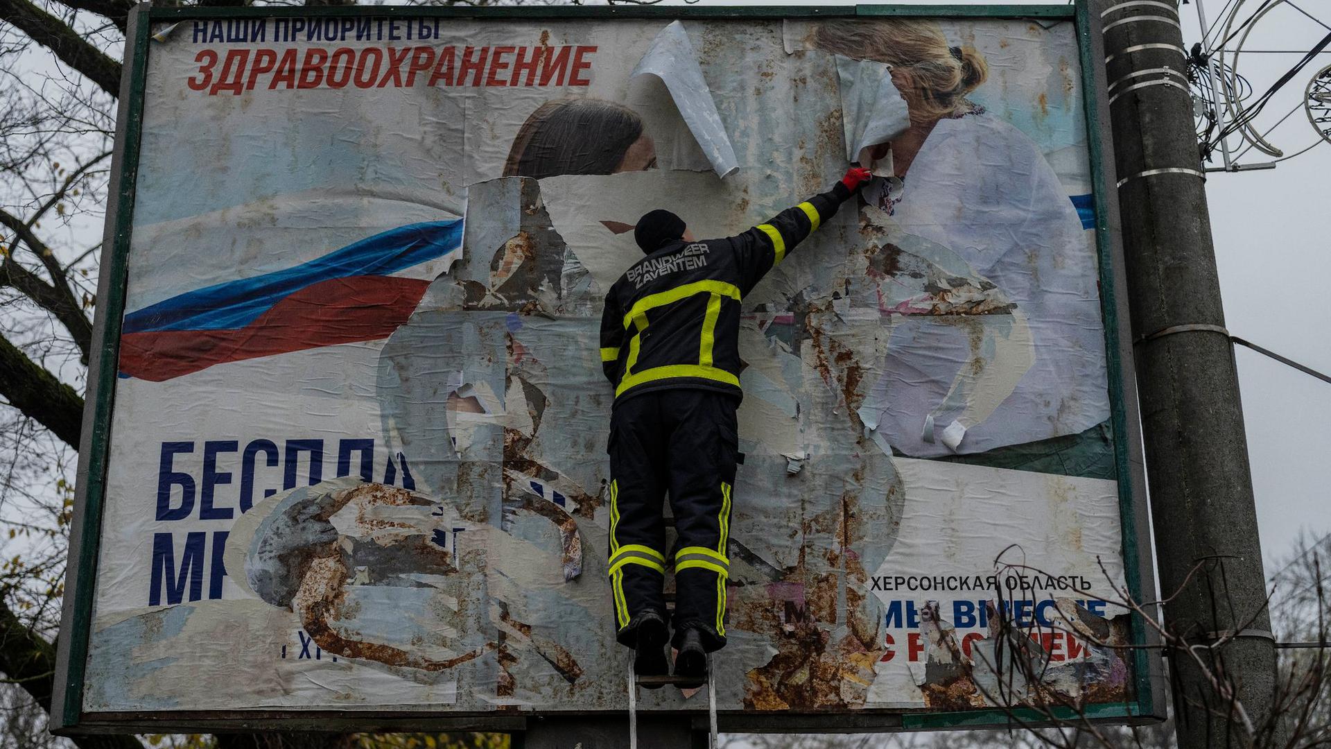 Ein ukrainischer Feuerwehrmann reißt in der kürzlich befreiten Stadt Cherson ein russisches Werbeplakat von einer Werbetafel.