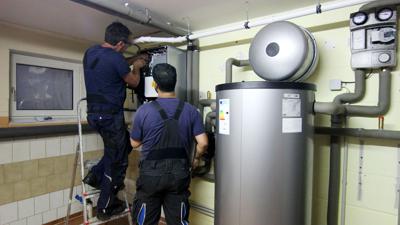 Zwei Handwerker stellen eine gerade installierte Wärmepumpe im Keller einer Doppelhaushälfte ein.