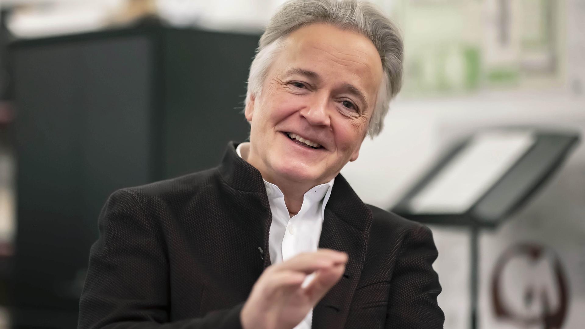 Der Rektor der Musikhochschule Karlsruhe lächelt in die Kamera und hebt dabei leicht die Hand